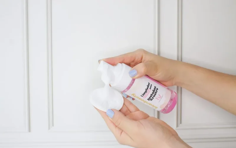 Close-up of hands applying skincare foam from a pink cosmetic bottle onto a cotton pad.