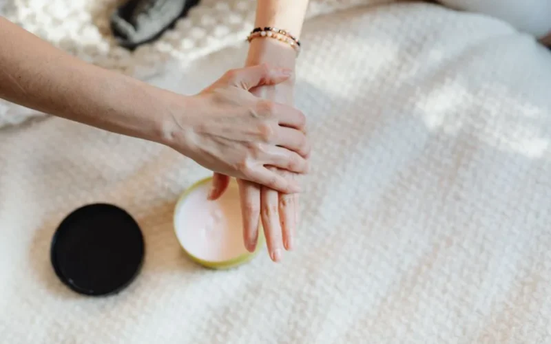 A close-up of a woman applying moisturizer on her hands with a cream container on a cozy bed.