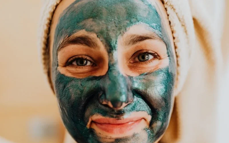 Close-up of a smiling woman with a green face mask, promoting skincare ritual.