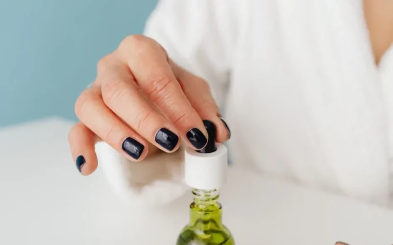 Hand holding a dropper over a glass bottle with green liquid, showcasing manicured nails.