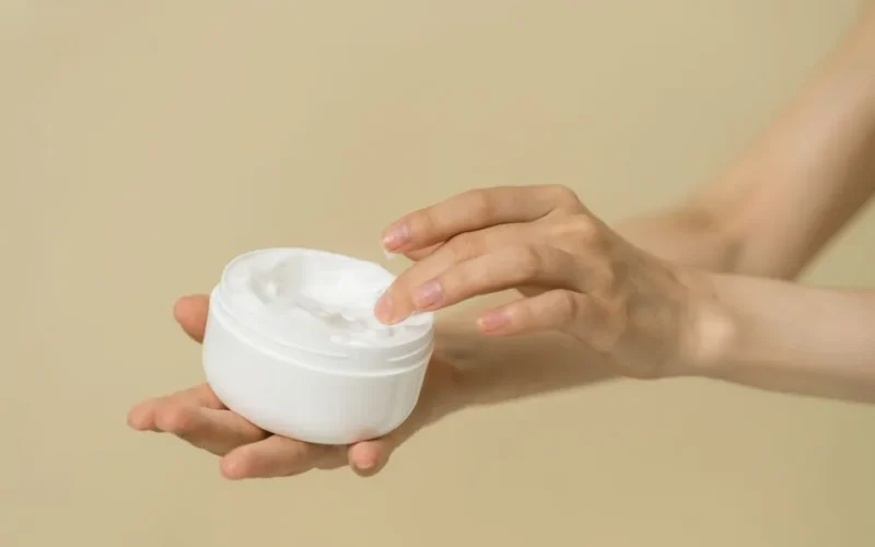 Close-up of female hands applying moisturizer from an open jar, highlighting skincare and self-care routine.