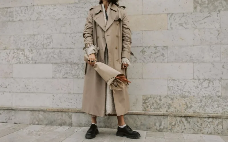 Stylish woman in beige trench coat holding a bouquet against a marble wall. Elegant urban fashion vibe.