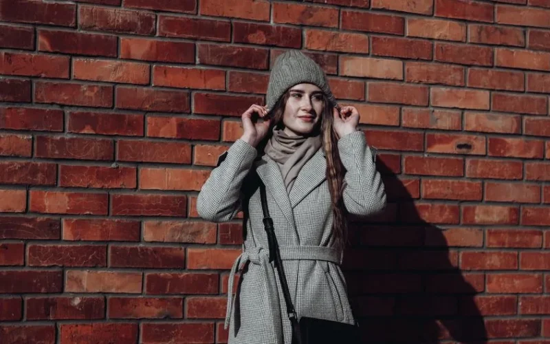Stylish woman in gray coat and beanie standing against a brick wall.