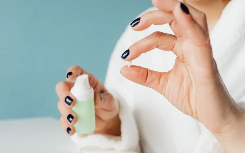 Close-up of a woman applying skincare cream with painted nails indoors.