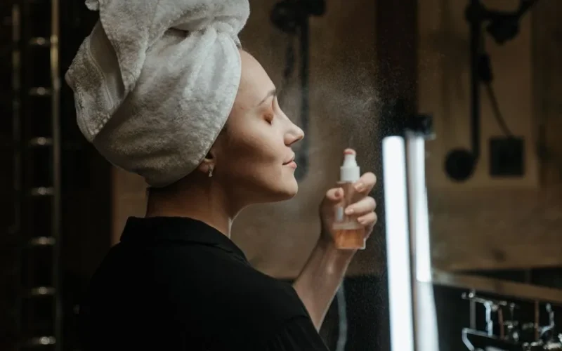 Adult woman with towel on head applies a refreshing facial mist in her skincare routine.