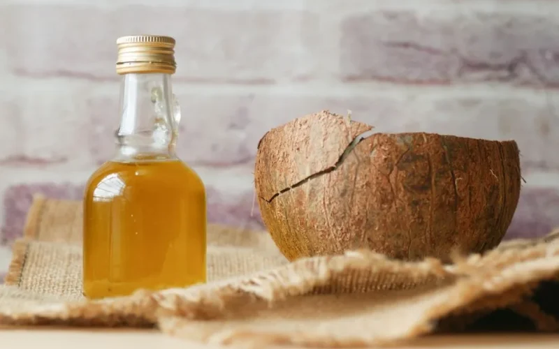 Bottle of coconut oil beside a cracked coconut shell on burlap fabric indoors.