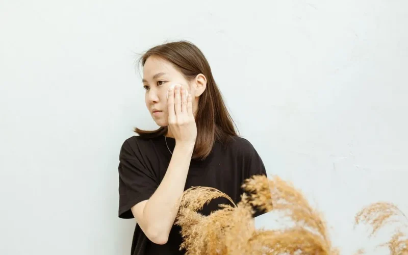 Asian woman applying skincare with a makeup remover pad, wearing a black shirt indoors.