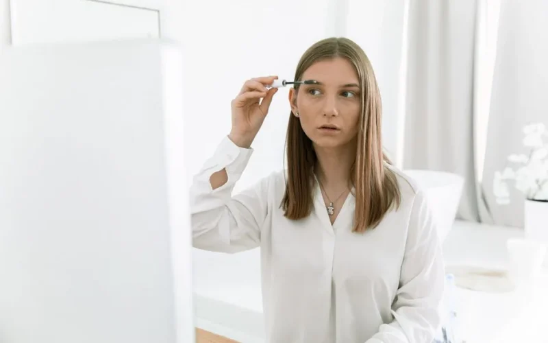 Young woman applying eyebrow makeup in a bright modern indoor bathroom setting.