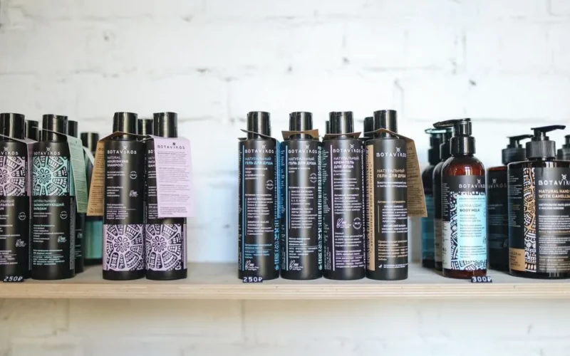 A variety of natural cosmetic bottles lined up on a wooden shelf against a white brick wall, suggesting eco-friendly beauty.