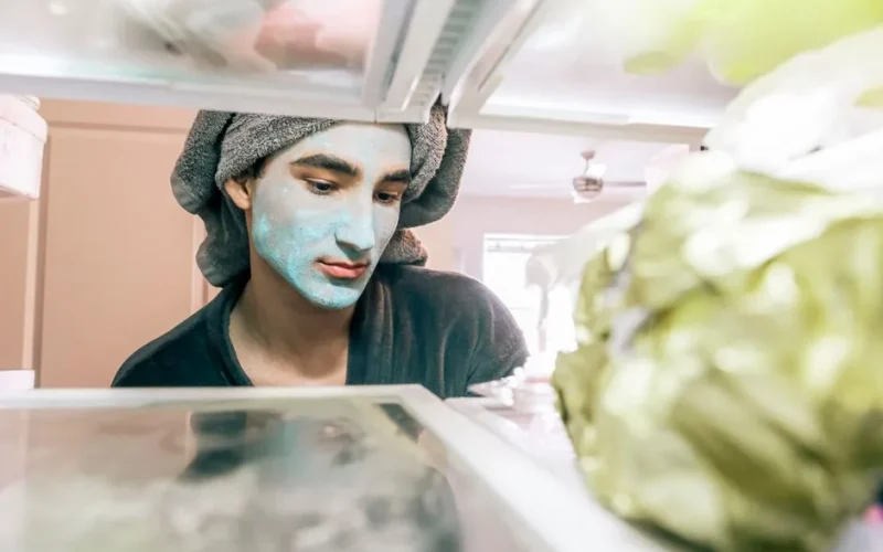 A man with a blue face mask looks into the refrigerator, towel wrapped on head, in a bright kitchen.