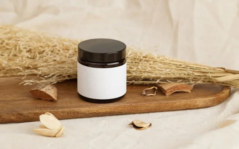 Natural cosmetic jar with blank label on a rustic wooden tray surrounded by dried herbs.