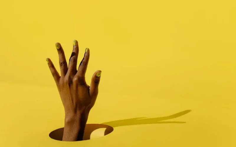 A hand with vibrant yellow nail art emerging through a hole against a bright yellow background.