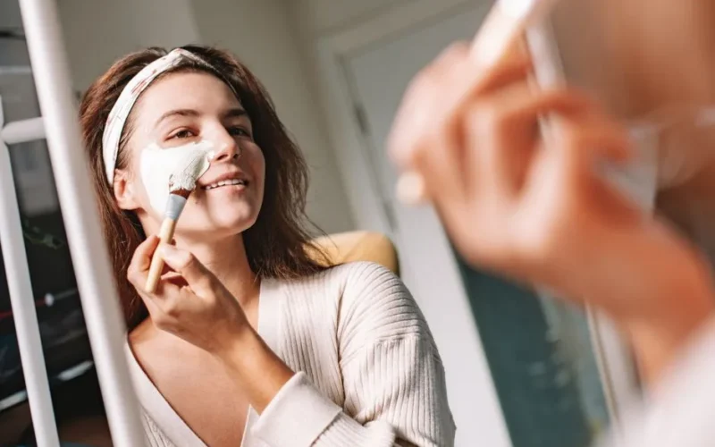 Woman enjoys a skincare routine, applying a facial mask with a brush in front of a mirror.