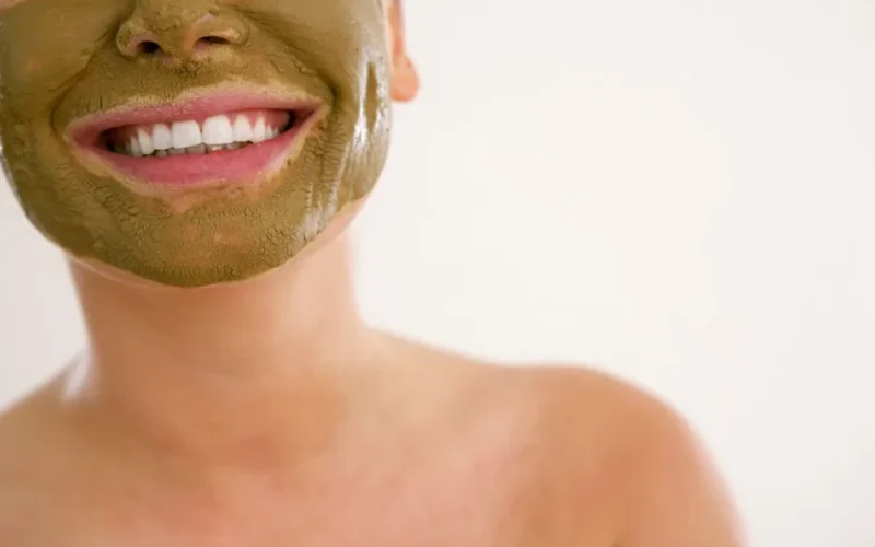 Close-up of a smiling woman applying a green cosmetic mask for skincare.