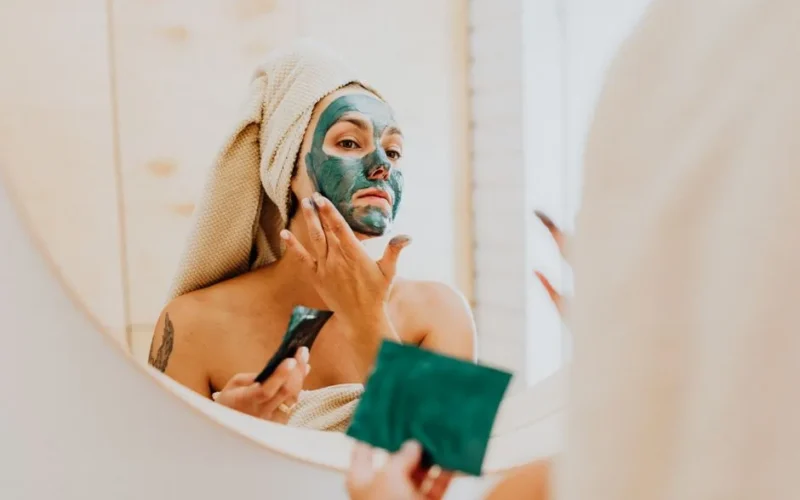 A woman applies a green clay face mask in a bathroom mirror, promoting self-care and skincare routines.