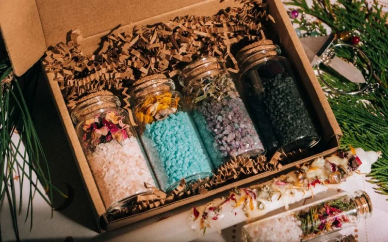 Top view of glass jars filled with colorful bath salt and aromatic dried herbs placed in cardboard box on table with green twigs