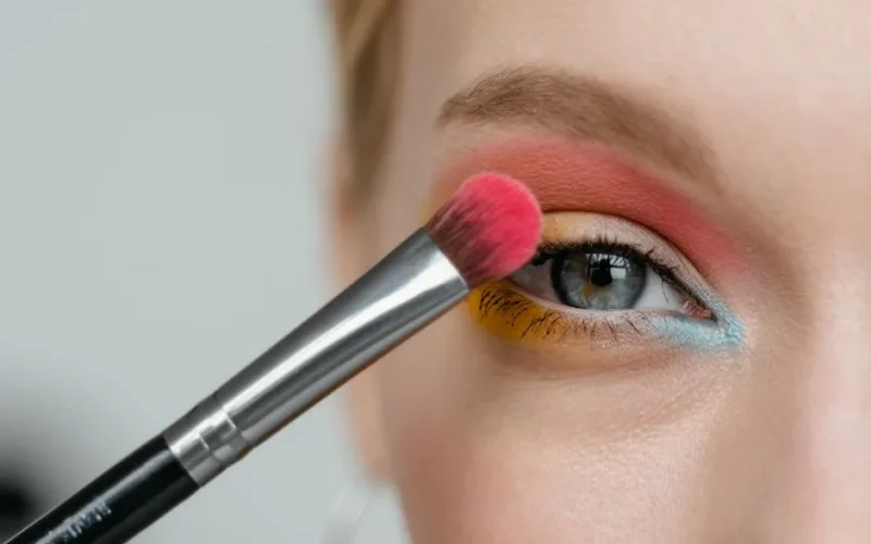 Close-up of a woman's eye with colorful makeup being applied with a brush.