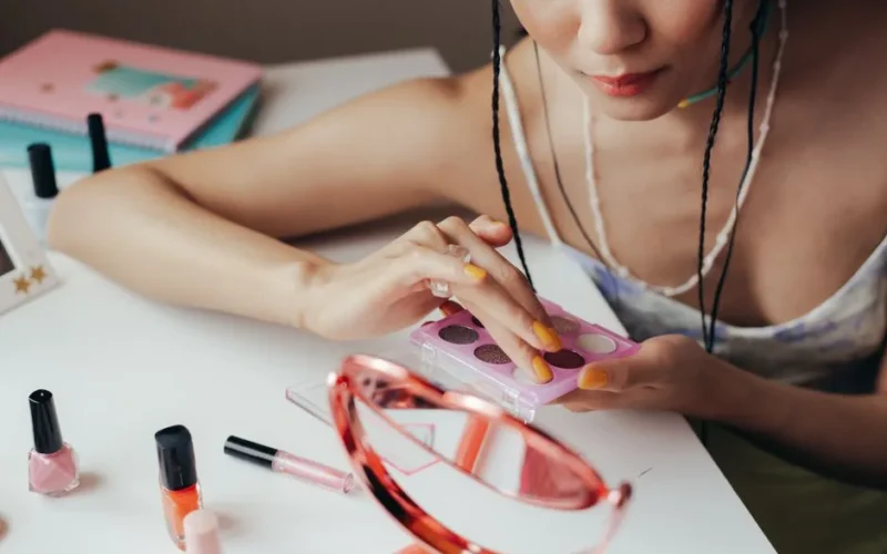 Woman using eyeshadow palette with cosmetics scattered on a table, reflecting in mirror.