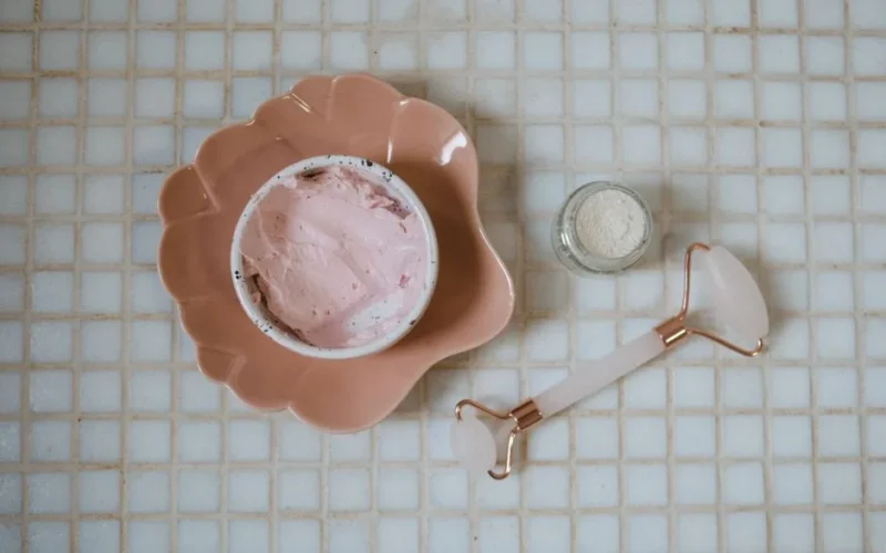 Close-up of spa essentials including pink cream, clay mask and jade roller on a tiled surface.