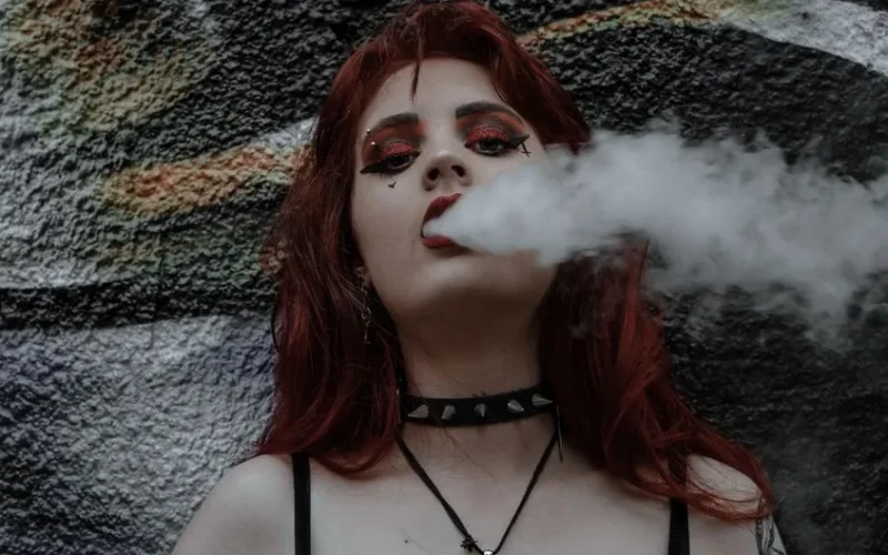 Portrait of a woman with red hair and bold eye makeup exhaling vapor against a graffiti wall.