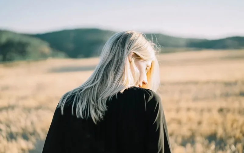 A woman with blonde hair stands in a sunlit field, creating a serene and tranquil atmosphere.