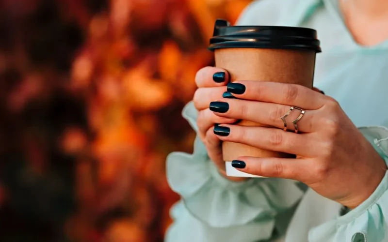 A woman's hands with dark nail polish holding a paper coffee cup against an autumn backdrop.