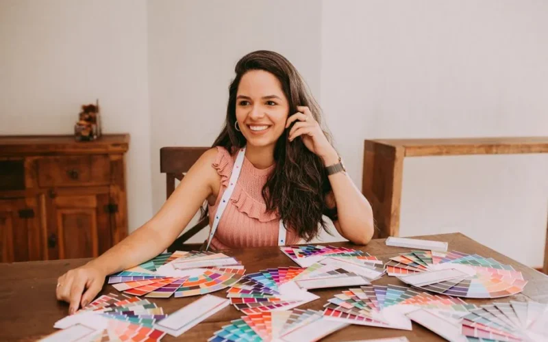 A woman with long hair, smiling while sitting at a table with various color swatches, suggesting creativity and design.