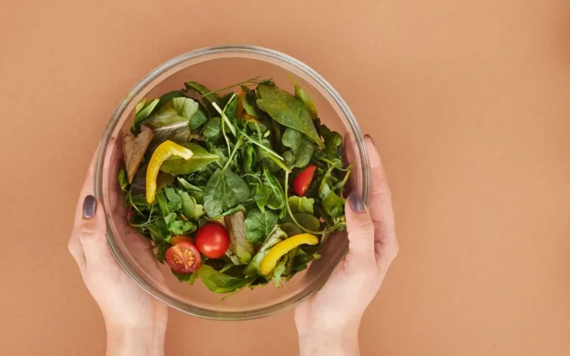 Healthy fresh salad bowl with cherry tomatoes and greens held by hands on a brown surface.