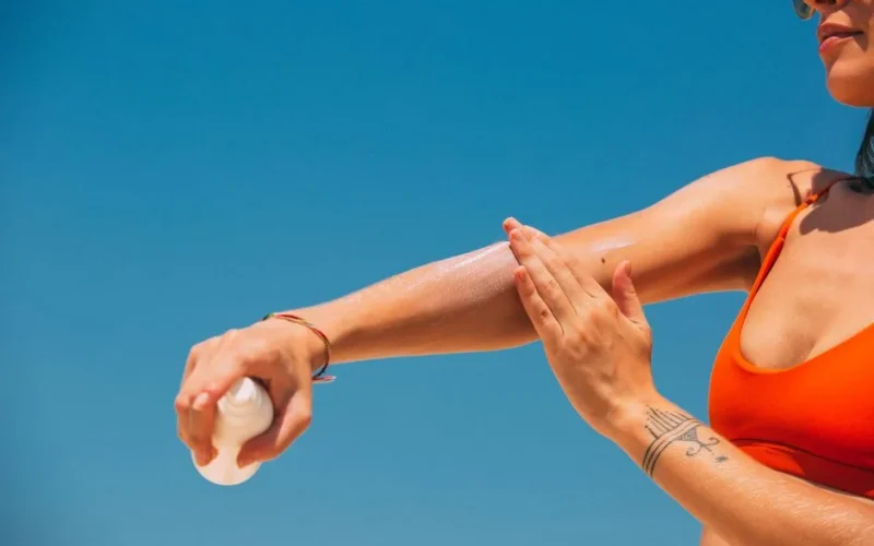 Close-up of a woman applying sunscreen on her arm under a clear blue sky at the beach.