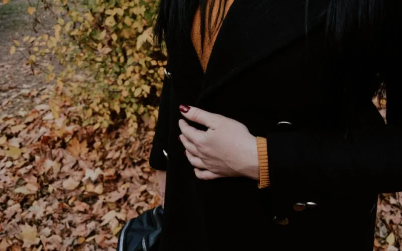 Woman with dark coat and painted nails standing near fall leaves in Kherson, Ukraine.