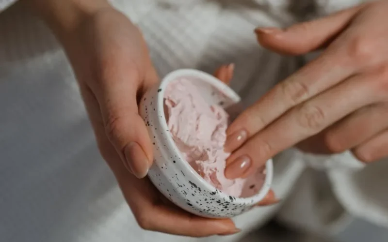 Close-up of hand applying pink moisturizing cream from a ceramic bowl for skincare routine.