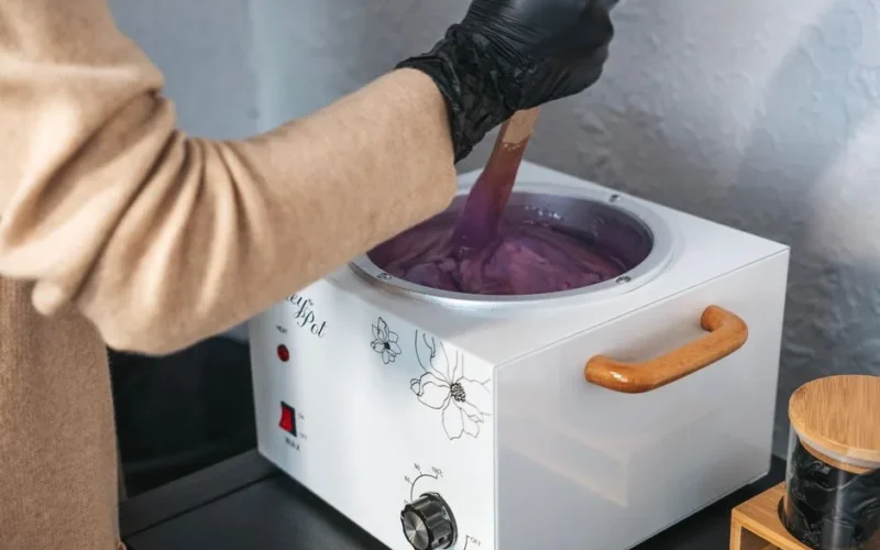An esthetician wearing gloves stirring purple wax in a wax pot indoors.