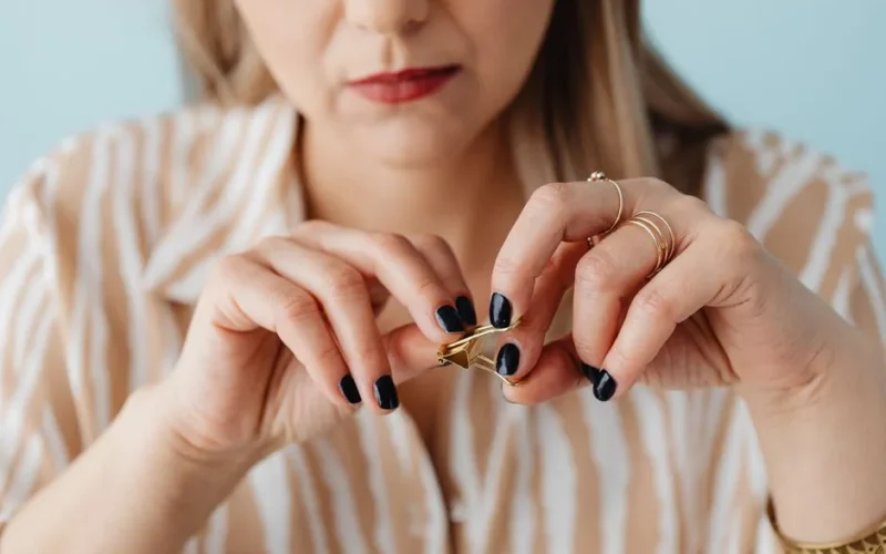 Close-up of a woman's hands fidgeting with a gold paperclip, showcasing painted nails and jewelry.