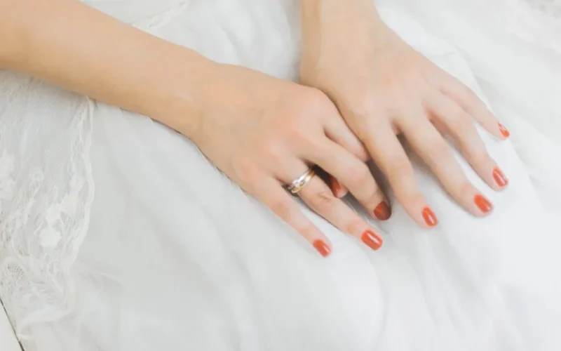 Close-up of a bride's hands with wedding ring and red nails on lace dress.