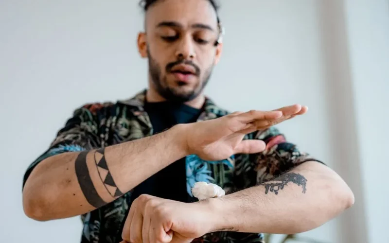 A man with tattoos applies cream to his forearm, highlighting tattoo care and skincare routines.