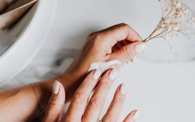 Close-up of manicured hands applying cream with a delicate floral accent on a marble surface.