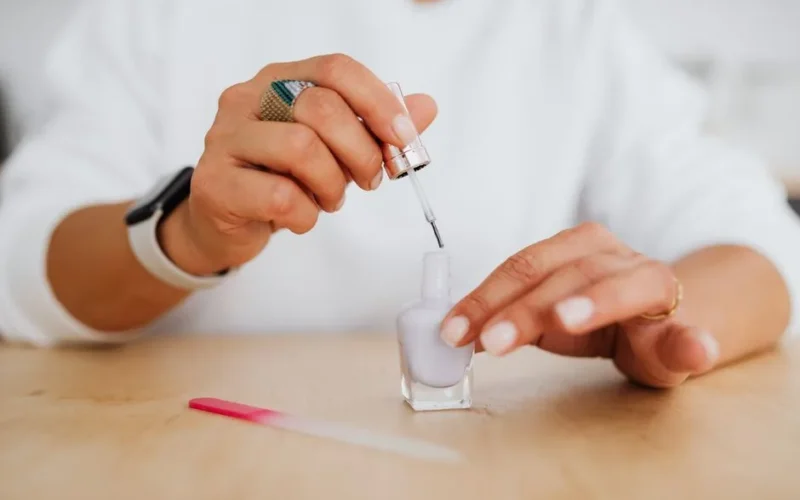 Close-up of hands applying lilac nail polish with a visible file on a wooden surface.