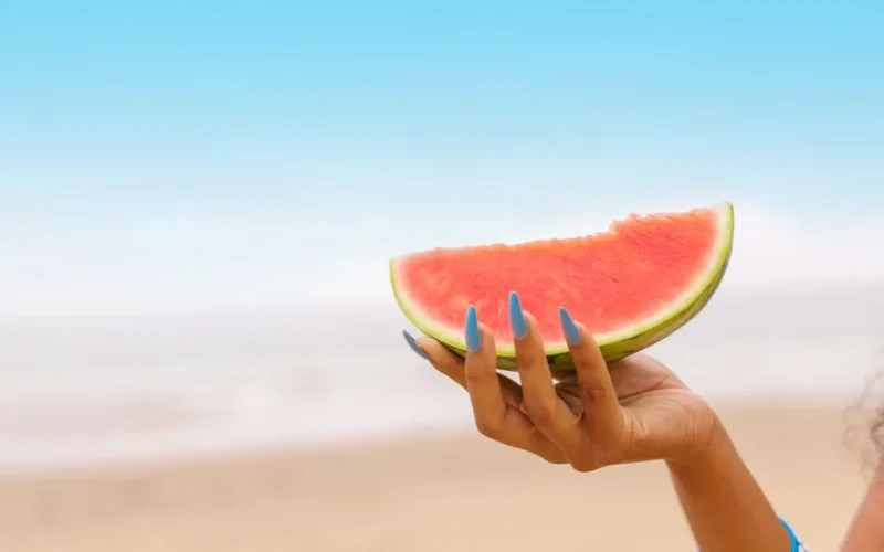 Vibrant watermelon slice held by a hand with blue nails on a sunny beach day.