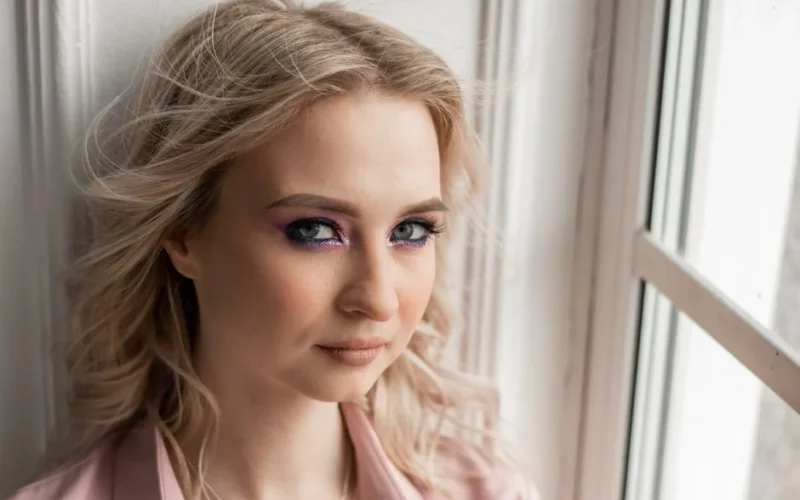 Close-up portrait of a blonde woman by a window with soft natural lighting.