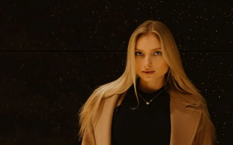 Fashion photography of a young woman with blonde hair against a black speckled background, wearing a stylish coat and necklace.
