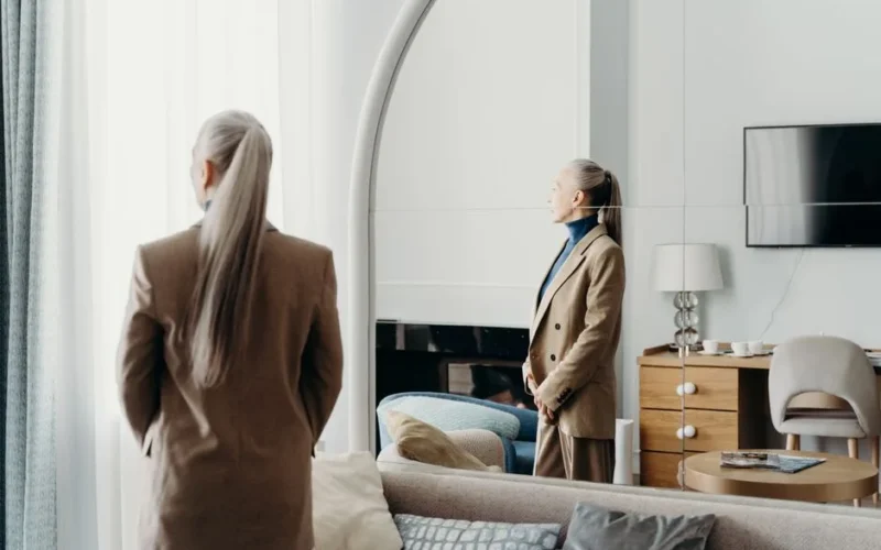 Sophisticated woman in a beige suit stands in an elegant room, viewing her reflection.