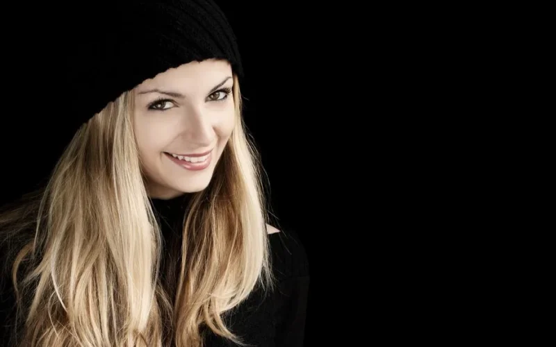 Beautiful woman with long blonde hair smiling in a black beanie against a dark background.