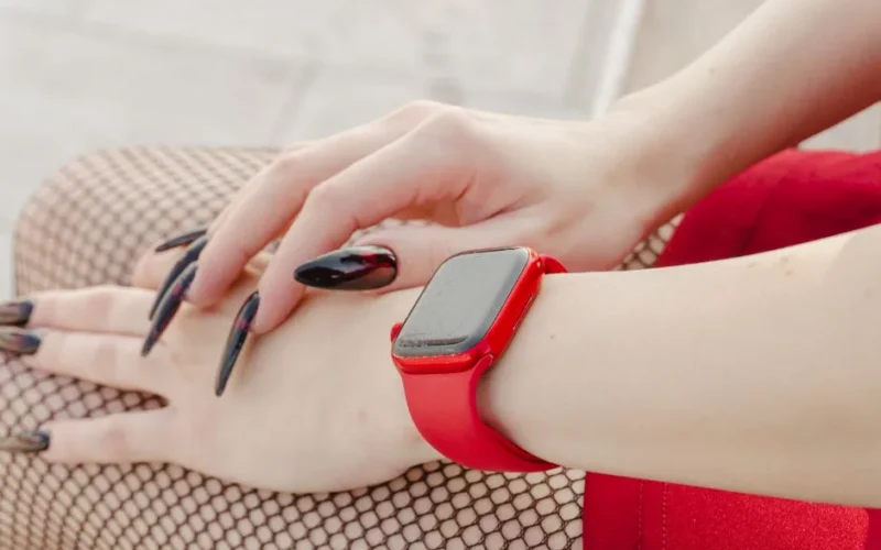 Close-up of a woman's hands with long nails and red smartwatch, showcasing modern fashion style.