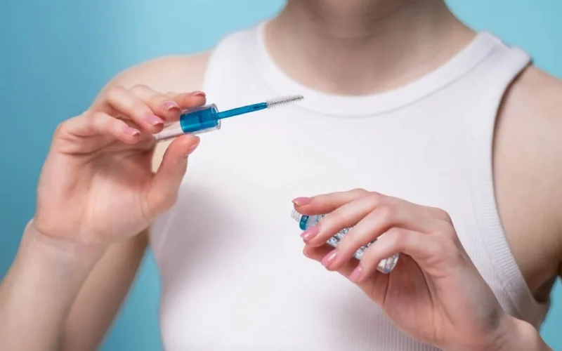 A close-up shot of a woman in a white top applying clear eyebrow gel with a blue wand.