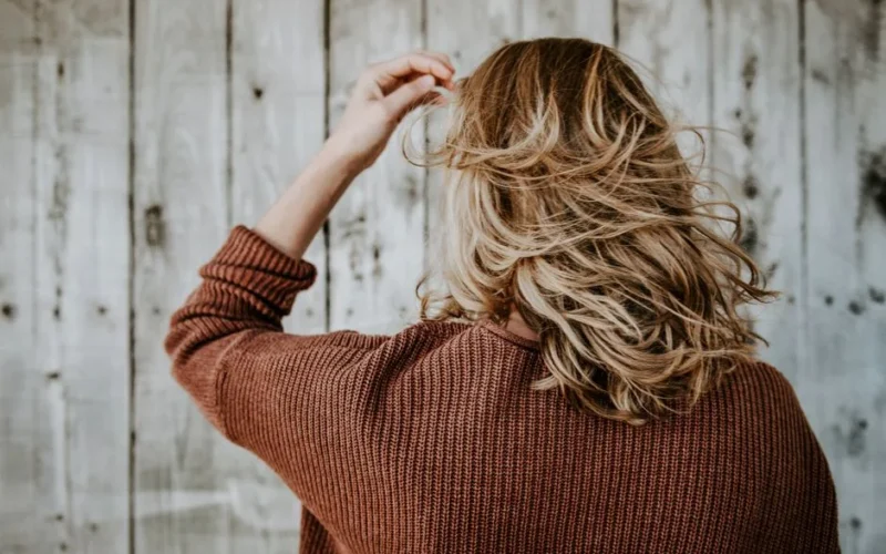 Rear view of a woman with blonde hair wearing a cozy sweater in front of rustic wooden wall.