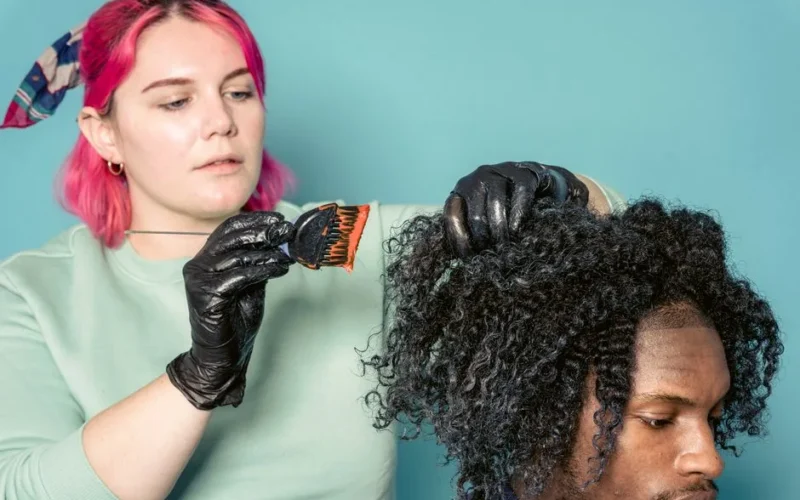 Serious woman with dyed hair applying paint on hair of African American model in studio against blue background