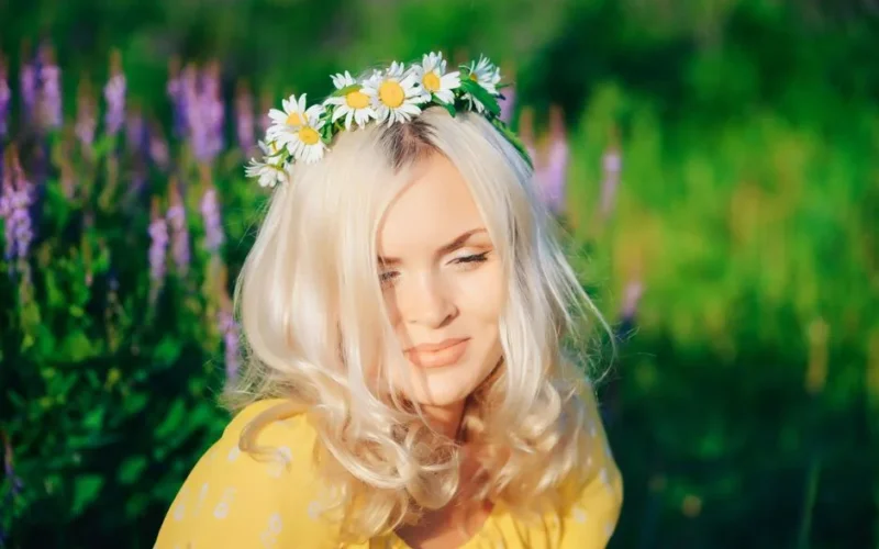 Blonde woman wearing a daisy crown enjoys a sunny day in a vibrant flower garden.