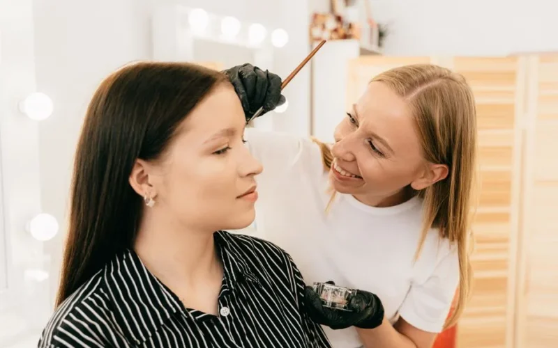 Makeup artist applying eyebrow tint on a smiling woman in a beauty salon.