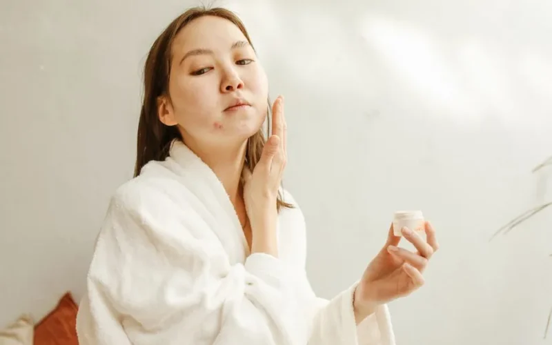 Close-up of an Asian woman in a bathrobe applying skincare cream as part of her beauty routine.
