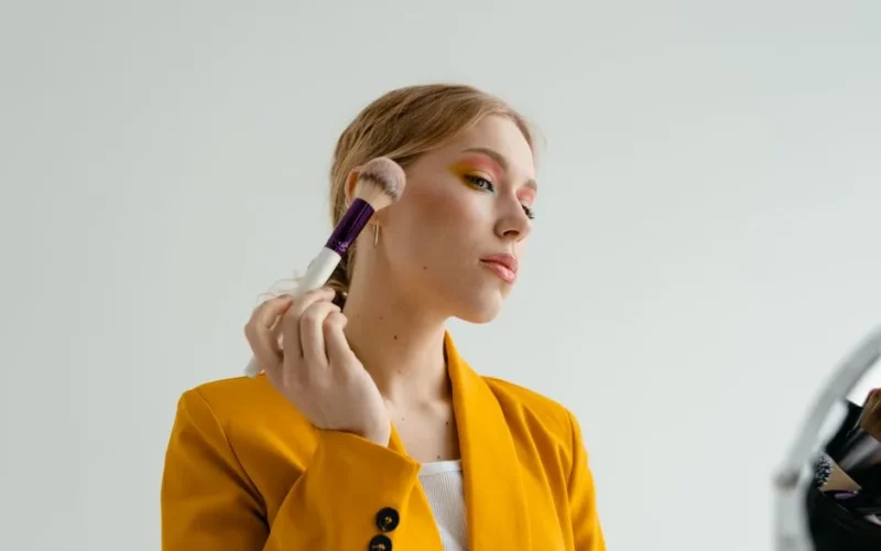 A woman in a yellow blazer applies makeup with a brush, showcasing a glamorous look.